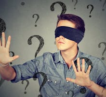 blindfolded man stretching his arms out walking through many question marks