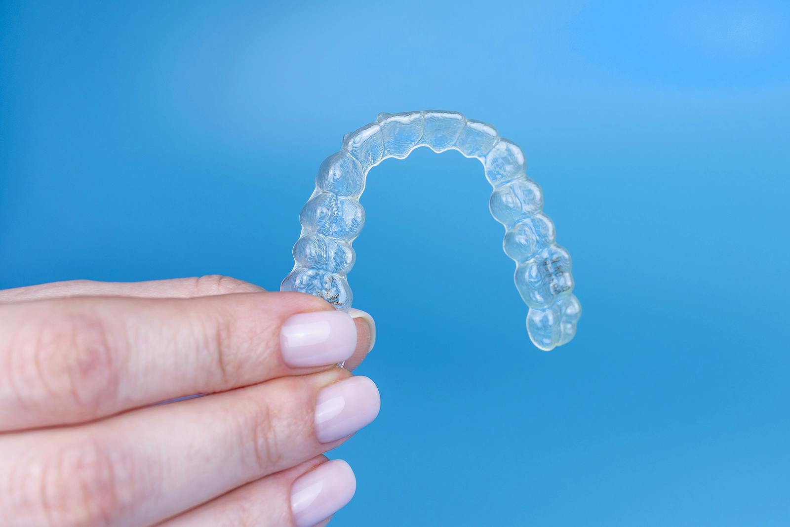 Hand holding removable invisible transparent aligner invisalign or plastic orthodontic braces retainer on blue background
