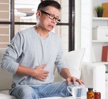 Mature 50s Asian man suffering heartburn or stomachache, pressing on stomach with painful expression, sitting on sofa at home, medicines on table