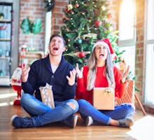 Two people opening packages by Christmas tree expressing holiday stress
