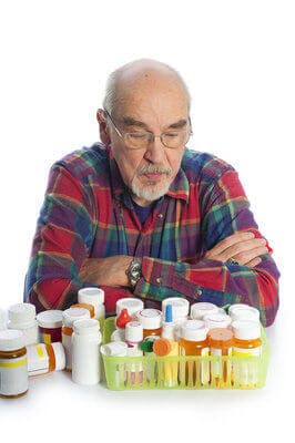 Senior Man, arms crossed, looks down at large assortment of prescription bottles pills
