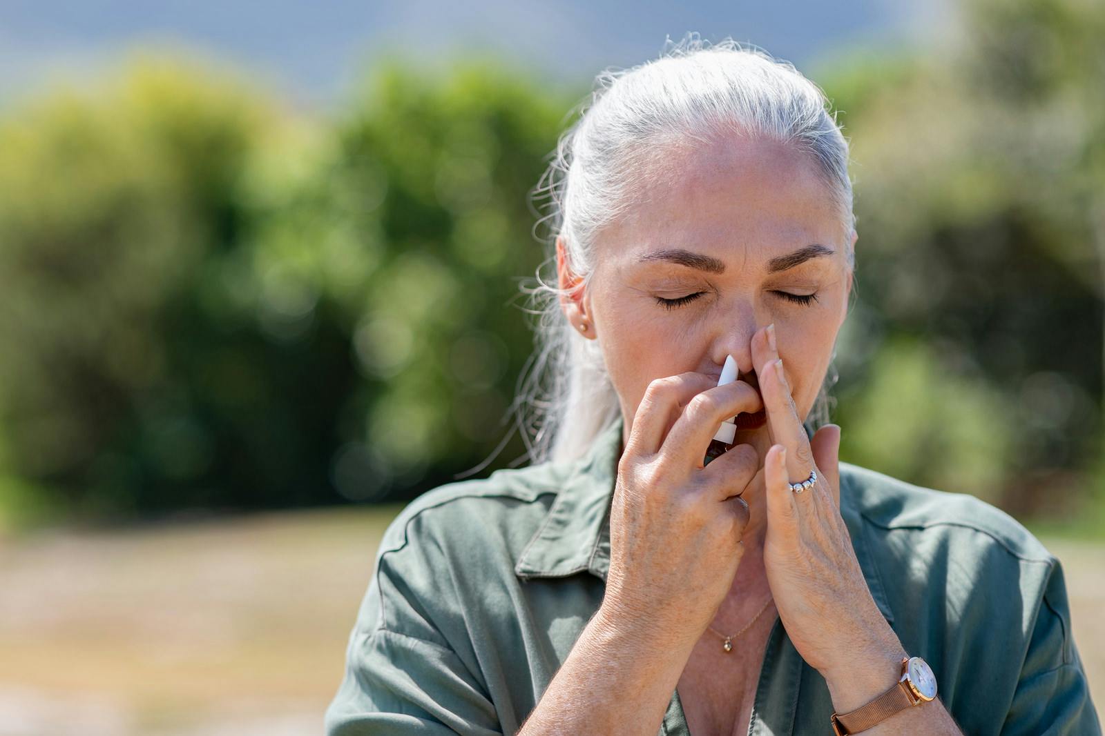 Senior woman having flu using nasal spray to help herself. Woman using a nasal spray at park for allergy. Ill mature woman with grey hair inhaling medicine against allergy.
