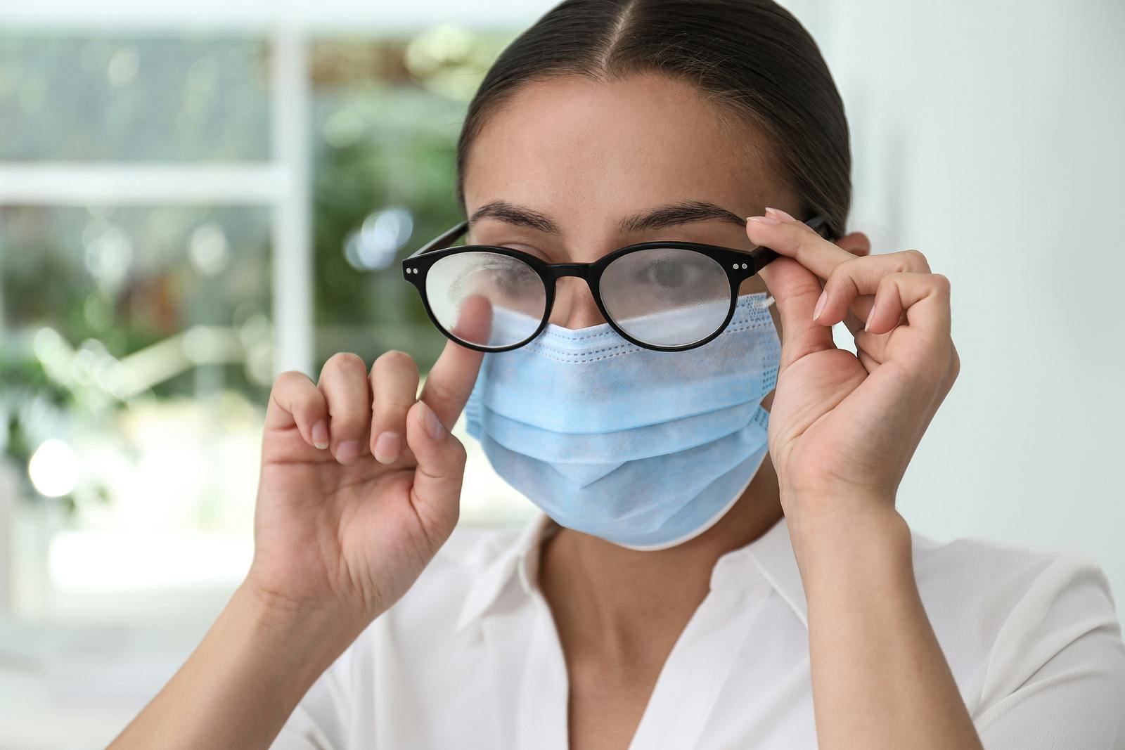 Woman wiping foggy glasses caused by wearing medical mask indoors, closeup
