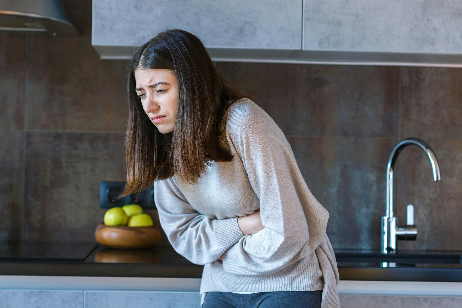 Young woman in grey clothes is holding hands on belly. Brunette girl is feeling bad and sick. Sudden onset of diarrhea, stomach ache, pancreatitis, appendicitis attack. Bad junk food concept.
